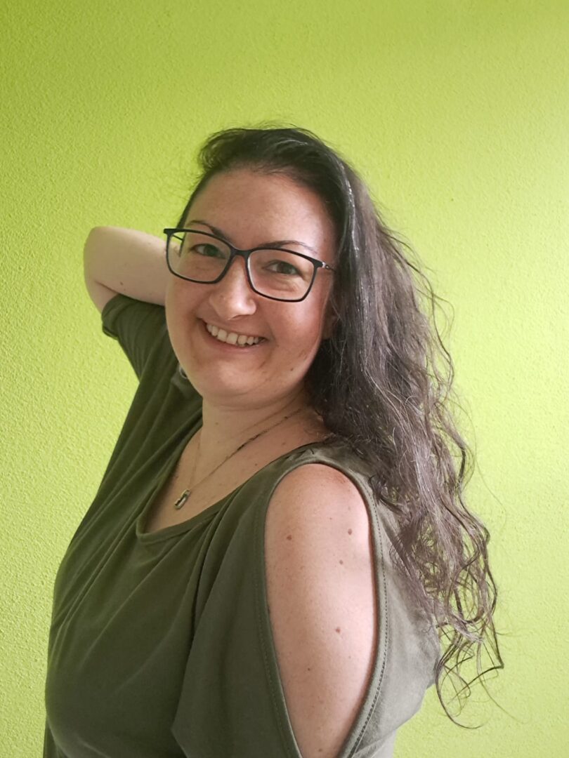 Nadja 2 Nadja lächelnd mit langen, lockigen Haaren und dunklen Brille steht vor einer hellgrünen Wand und blickt freundlich in die Kamera.