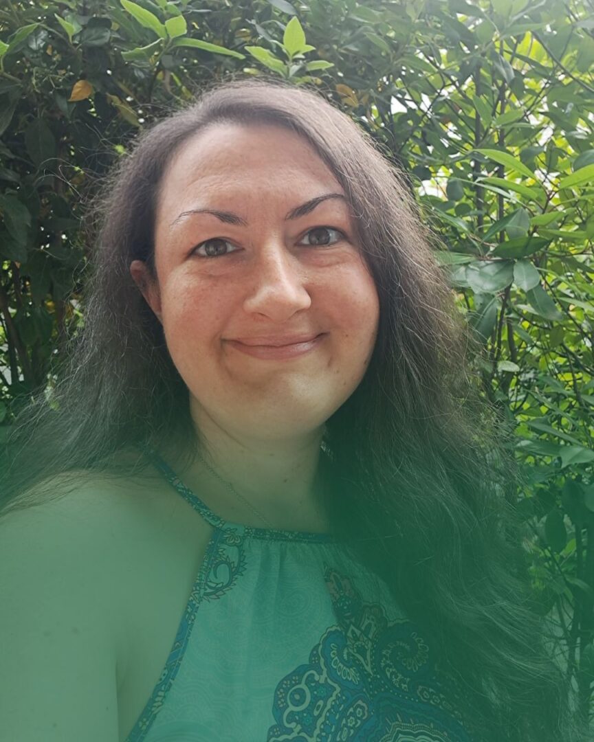 Nadja Nadja mit langen, dunklen Haaren lächelt in die Kamera. Sie trägt ein ärmelloses Oberteil mit blauem Muster. Im Hintergrund sind grüne Blätter und Pflanzen.